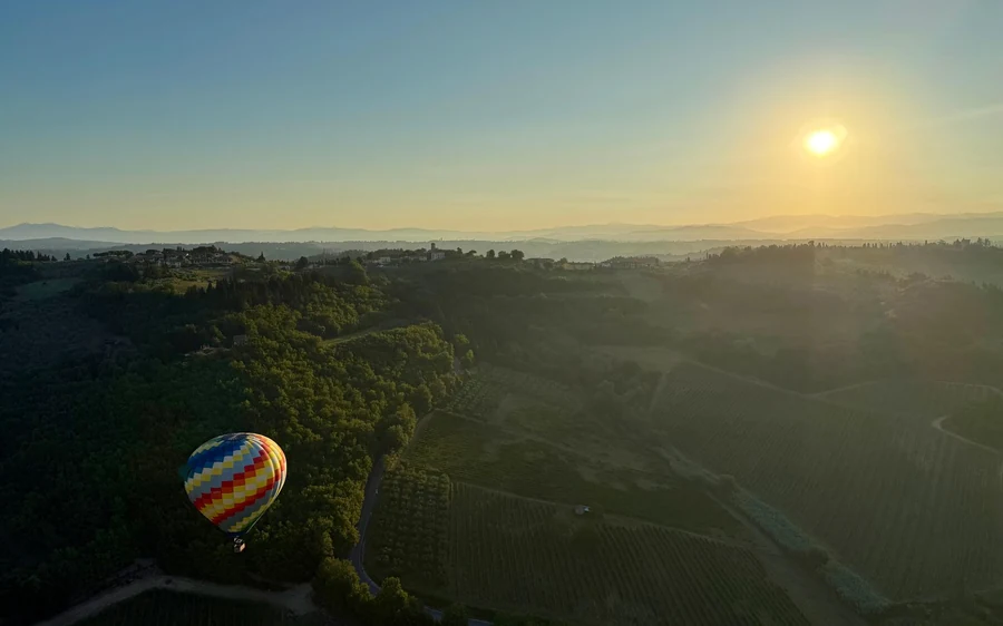 A striped balloon drifting low over wooded slopes and vineyards with the sun just up