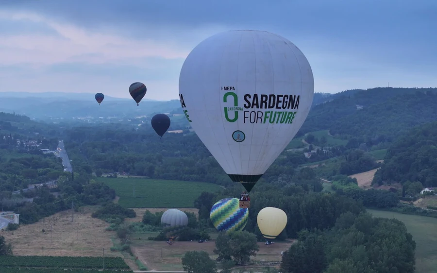 A large white balloon in flight above wooded hills, with three more balloons further off